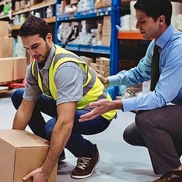 A trainer coaching a warehouse worker in a hi-vis vest on correct manual handling technique while lifting a box